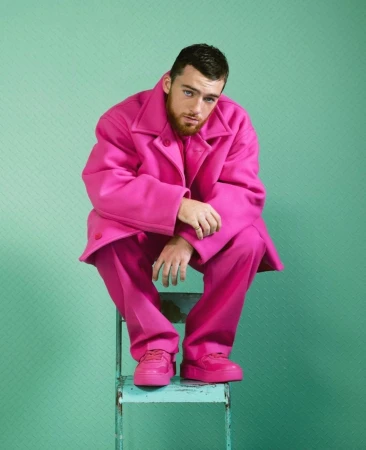 Portrait of a man in a bright pink suit sitting on a blue metal step stool against a light green textured background, eye-level shot.