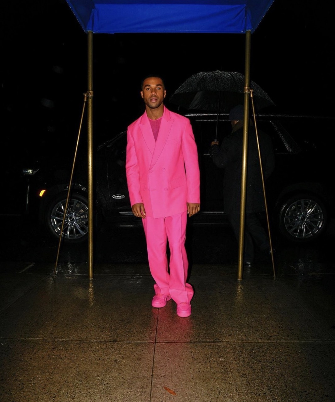 A man in a vibrant pink suit stands outdoors on a wet surface, illuminated by artificial light, with a black SUV in the background.