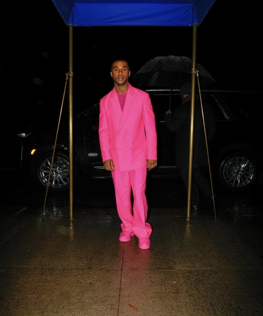 A man in a vibrant pink suit stands outdoors on a wet surface, illuminated by artificial light, with a black SUV in the background.