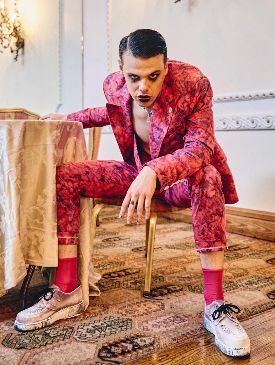 Young adult male in a patterned pink suit sitting on a chair in an indoor environment with neutral backgrounds and unique styling.