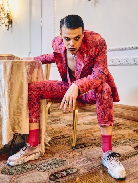 Young adult male in a patterned pink suit sitting on a chair in an indoor environment with neutral backgrounds and unique styling.