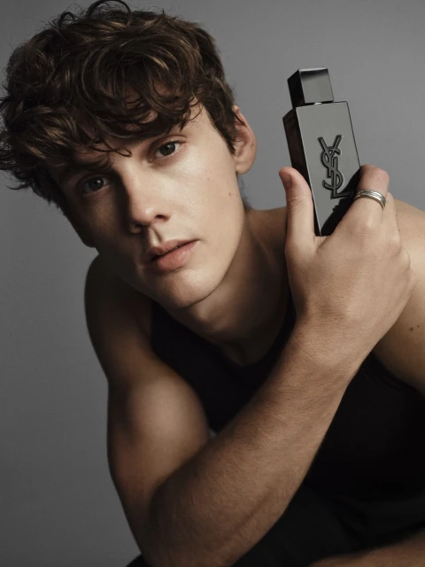 Portrait of a fair-skinned young adult male holding a black bottle of YSL cologne against a solid grey backdrop in a photo studio.