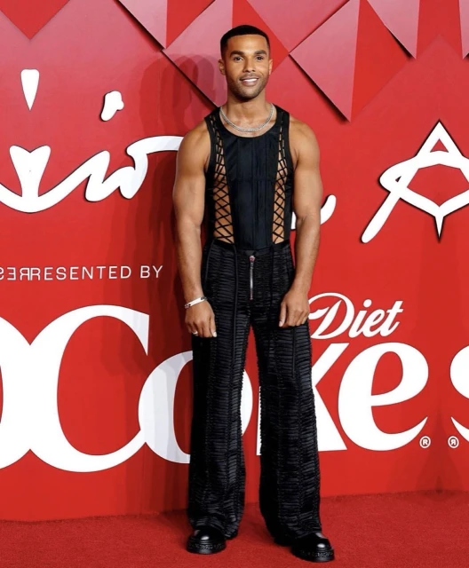Man poses on red carpet in black textured outfit against a red backdrop with white Diet Coke logos and geometric patterns.
