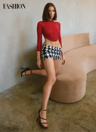 Fashionable young woman poses indoors wearing a red long-sleeved crop top, patterned miniskirt, and black strappy high-heeled sandals.