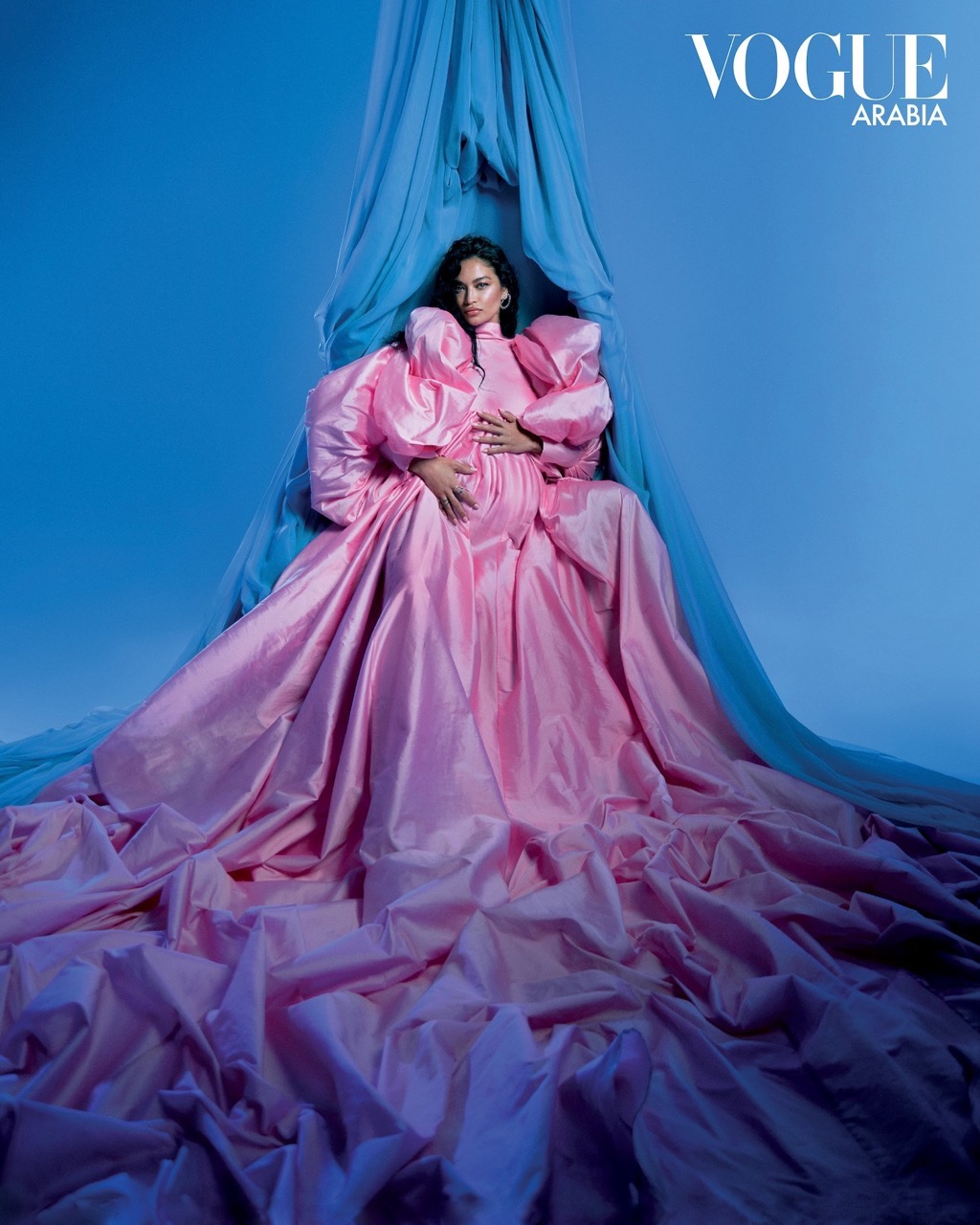 A pregnant woman in an elaborate pink gown poses in a blue-lit studio. Vogue Arabia logo visible. Conceptual fashion photography.