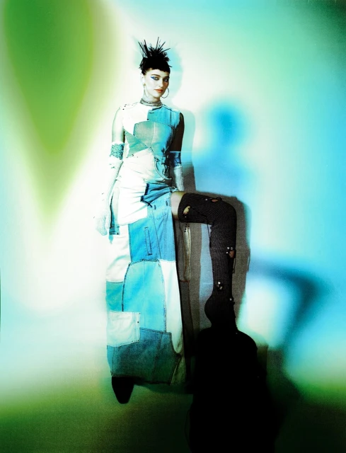 A stylish adult female poses with a leg raised in knee-high hosiery. She wears a patchwork dress in front of a bright blue and green backdrop.