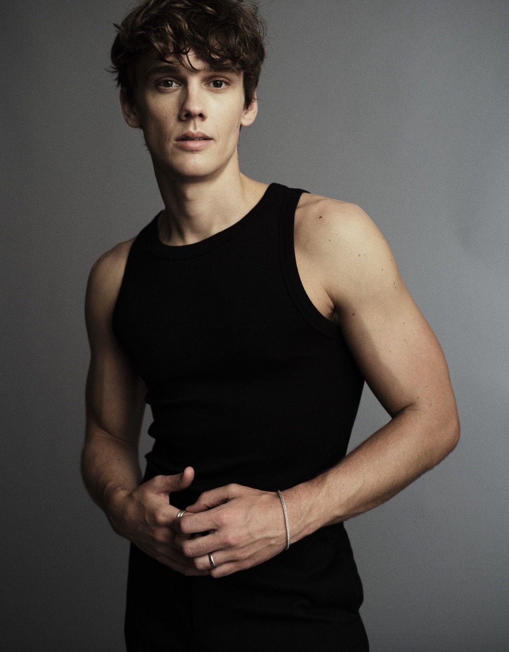 Portrait of a fit young adult male wearing a black tank top in a photo studio. He has brown curly hair and fair skin and is wearing jewelry.