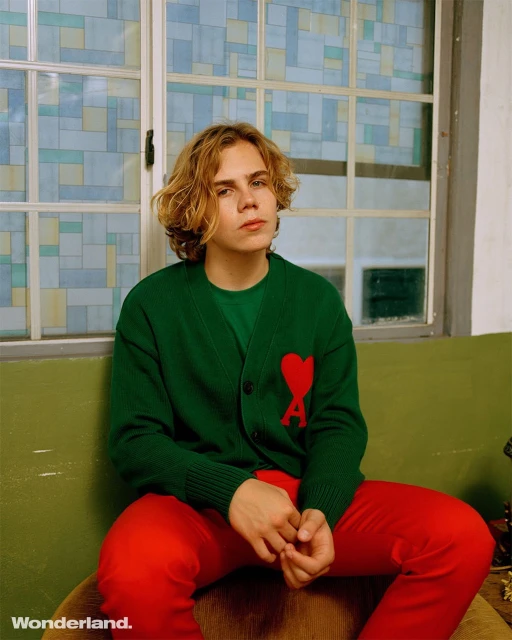 Portrait of a blond male in a green cardigan with a red heart, red trousers, seated indoors near a window with geometric stained glass.