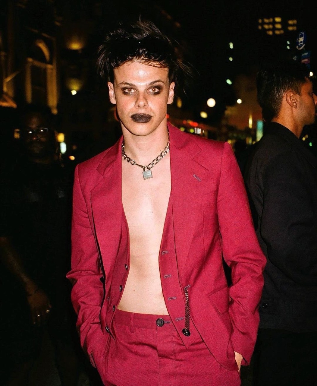 Young man in a red suit with dark makeup and chain necklace, standing outdoors at night with blurry figures in the background.