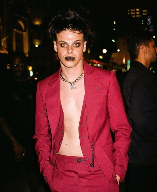 Young man in a red suit with dark makeup and chain necklace, standing outdoors at night with blurry figures in the background.