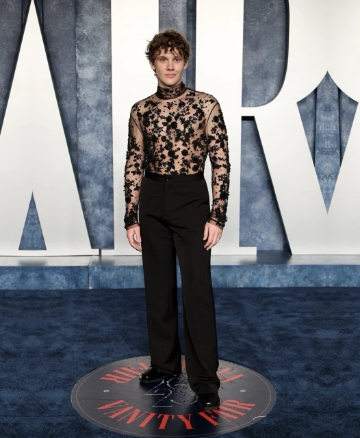 Man in a sheer floral-patterned top and black pants poses on a Vanity Fair carpet against a white branded backdrop.