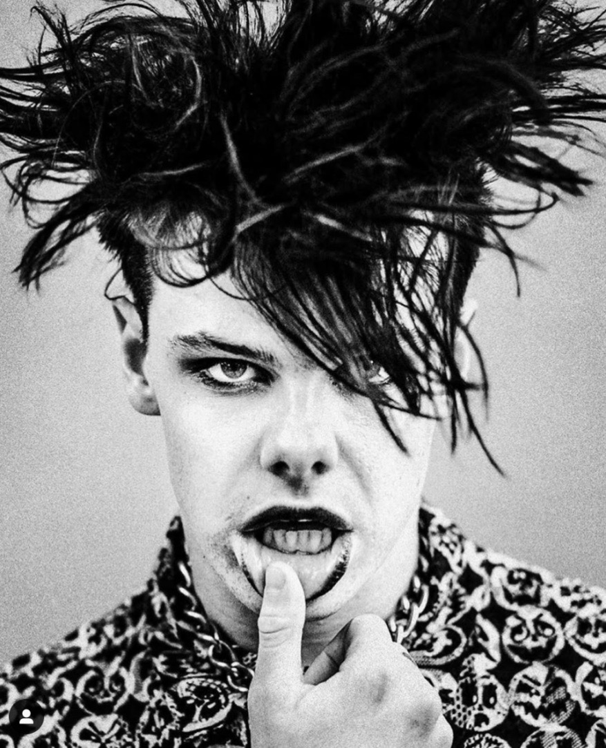 Monochrome portrait of a person with dramatic black makeup, wild dark hair, and a patterned top posing indoors.