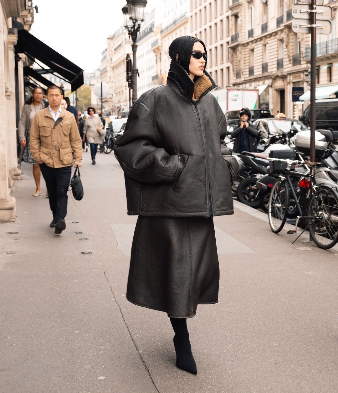 Woman in black coat walks on city street. She wears dark sunglasses, a beanie, skirt, and heeled shoes. Buildings and traffic in background.