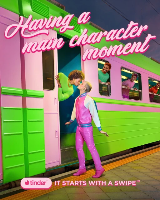 Two women kiss near a pink and green train, as 3 men watch from the train windows; the logo for Tinder is visible.