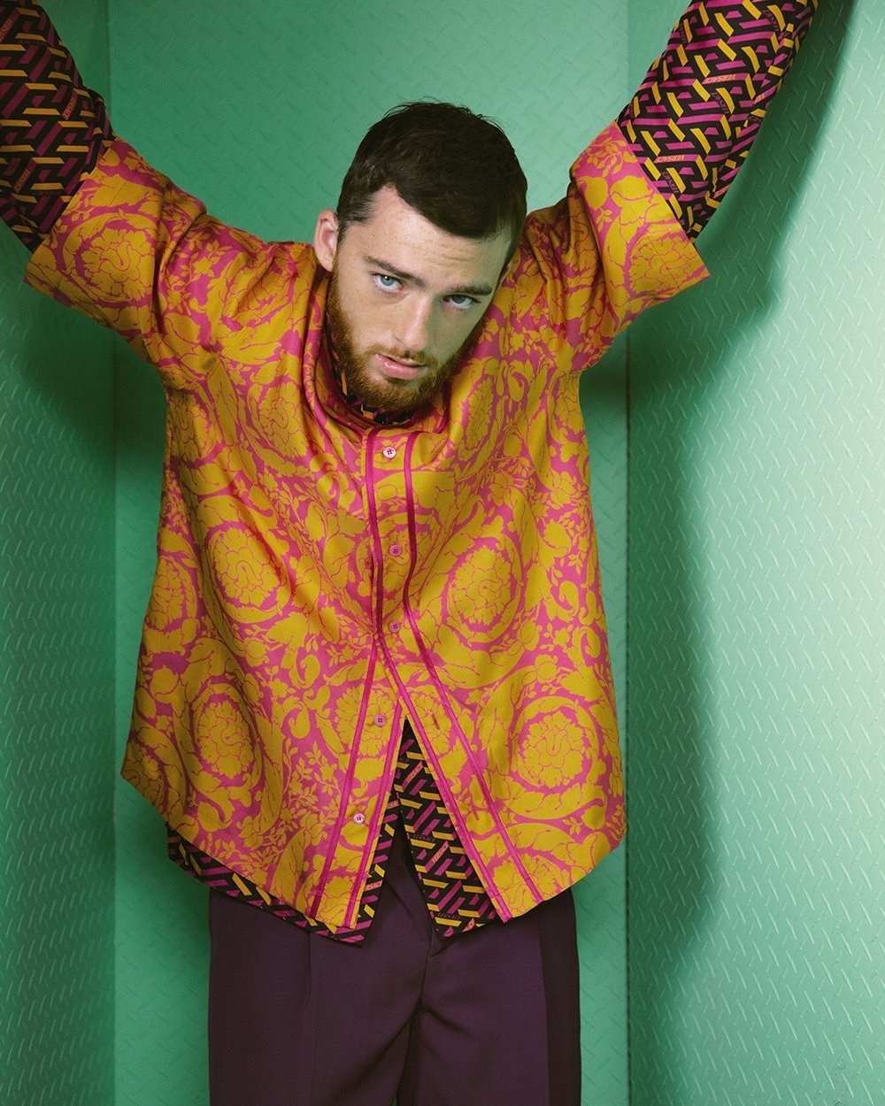 Fashion shot of a young adult male posing in a green-walled corner wearing a gold and pink patterned shirt and purple pants.