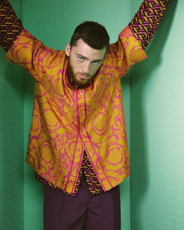 Fashion shot of a young adult male posing in a green-walled corner wearing a gold and pink patterned shirt and purple pants.