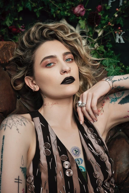 Portrait of a fair-skinned adult female with blonde curly hair, dark lipstick, visible tattoos, button vest against a floral background.