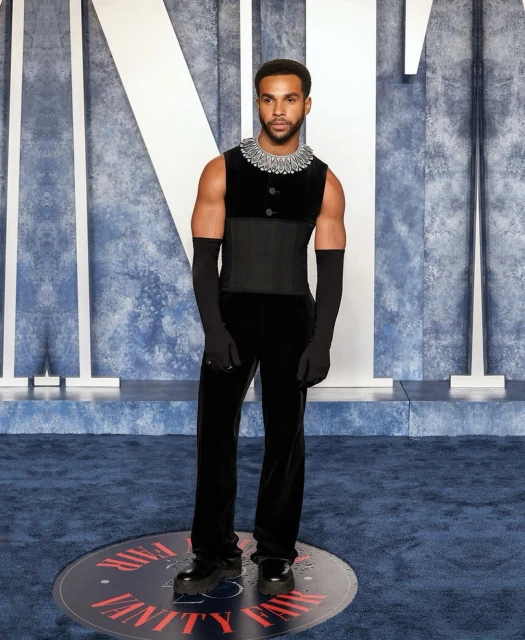 Man in black velvet sleeveless top, corset, and long gloves posing against a blue backdrop, on Vanity Fair logo, celebrity event.