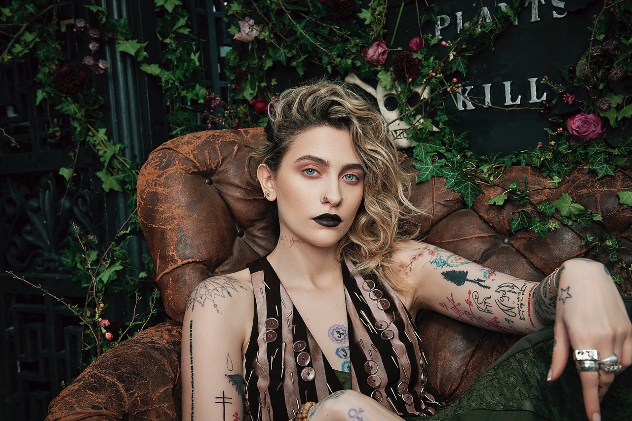 Close-up shot of Paris Jackson seated in an antique leather chair, adorned with arm tattoos, dark lipstick, and blonde curly hair.