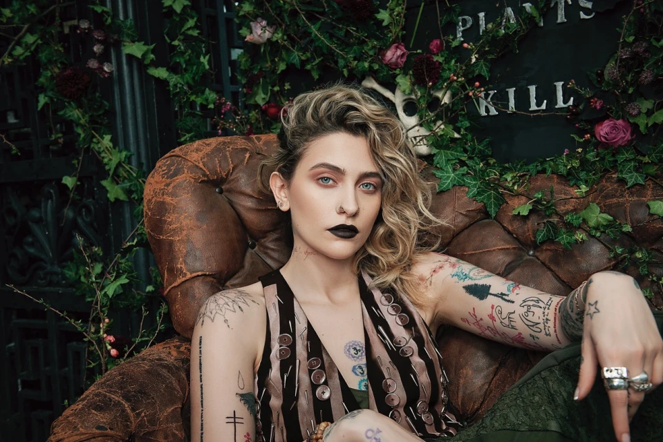 Close-up shot of Paris Jackson seated in an antique leather chair, adorned with arm tattoos, dark lipstick, and blonde curly hair.
