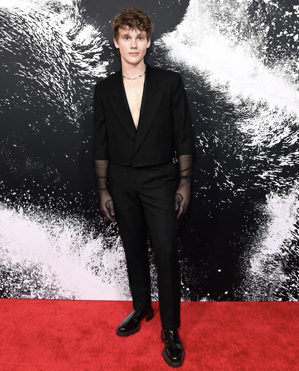 Full-length portrait of a young adult male with curly reddish-brown hair on a red carpet in a black, open blazer and black pants.