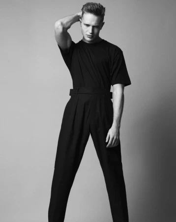 Man wearing a dark t-shirt and high-waisted belted trousers posing against a grey seamless studio backdrop.