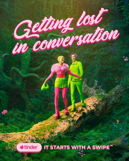 Two fashion models stand on a log in a whimsical forest for a Tinder advertisement, both wearing bright, retro-inspired clothing.