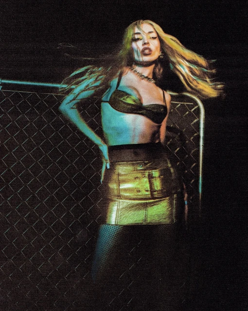 A woman poses in a metallic bra and skirt, highlighted by dramatic lighting against a chain-link fence background, creating an edgy fashion shot.