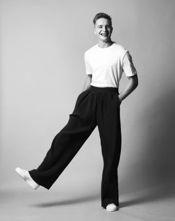 A smiling young man in a white t-shirt and wide black trousers posing with one foot lifted against a light grey seamless backdrop.