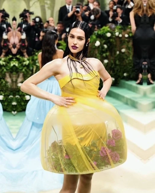 A woman poses in a yellow dress with visible roses and foliage in its skirt, at a crowded indoor event with photographers.