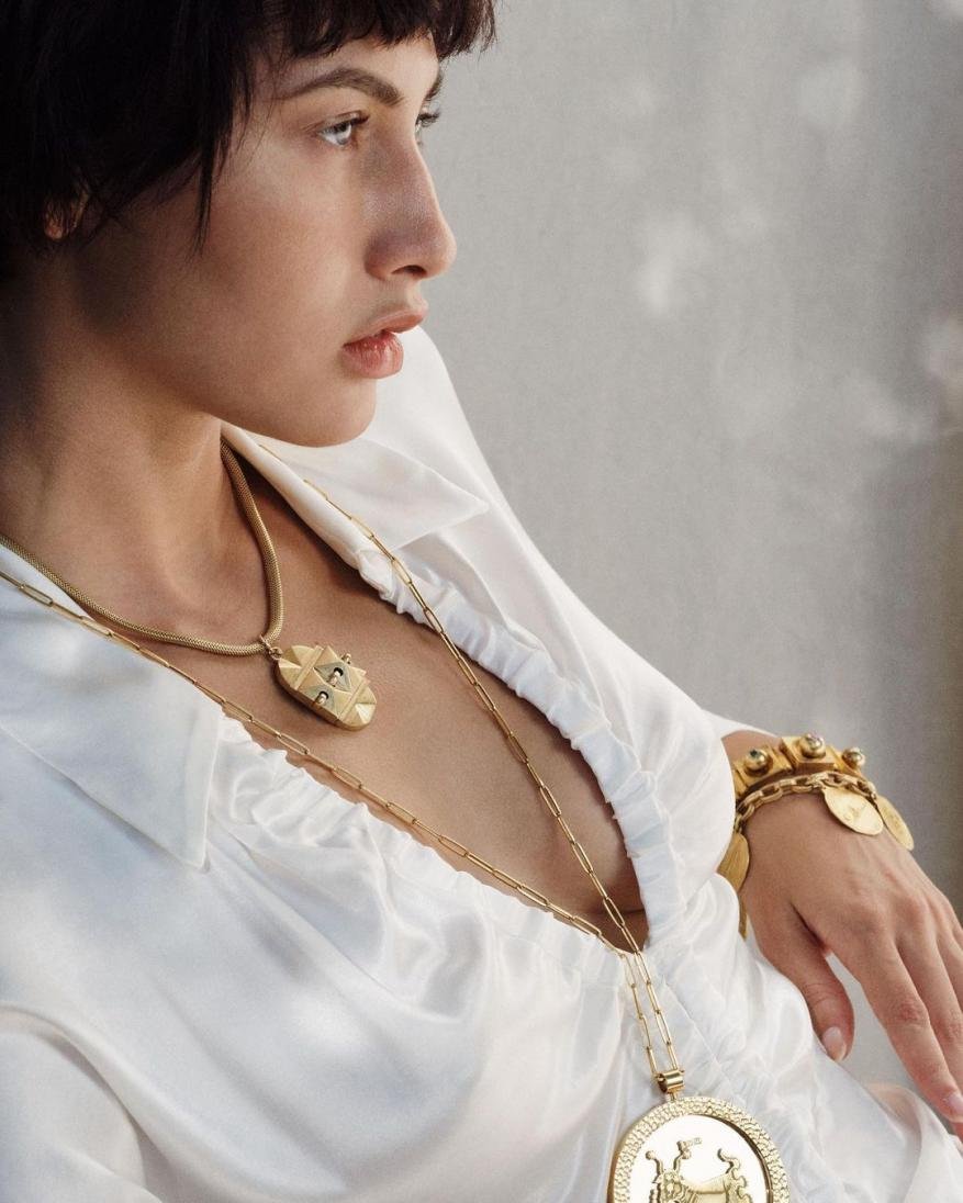 Woman in white shirt wearing gold necklaces and bracelet in soft, natural light showcasing fashion jewellery in a beauty-style setting.