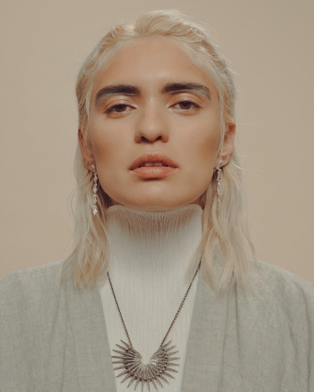 Close-up portrait of a woman with bleached blonde hair wearing a white turtleneck, grey jacket and necklace against a tan backdrop.