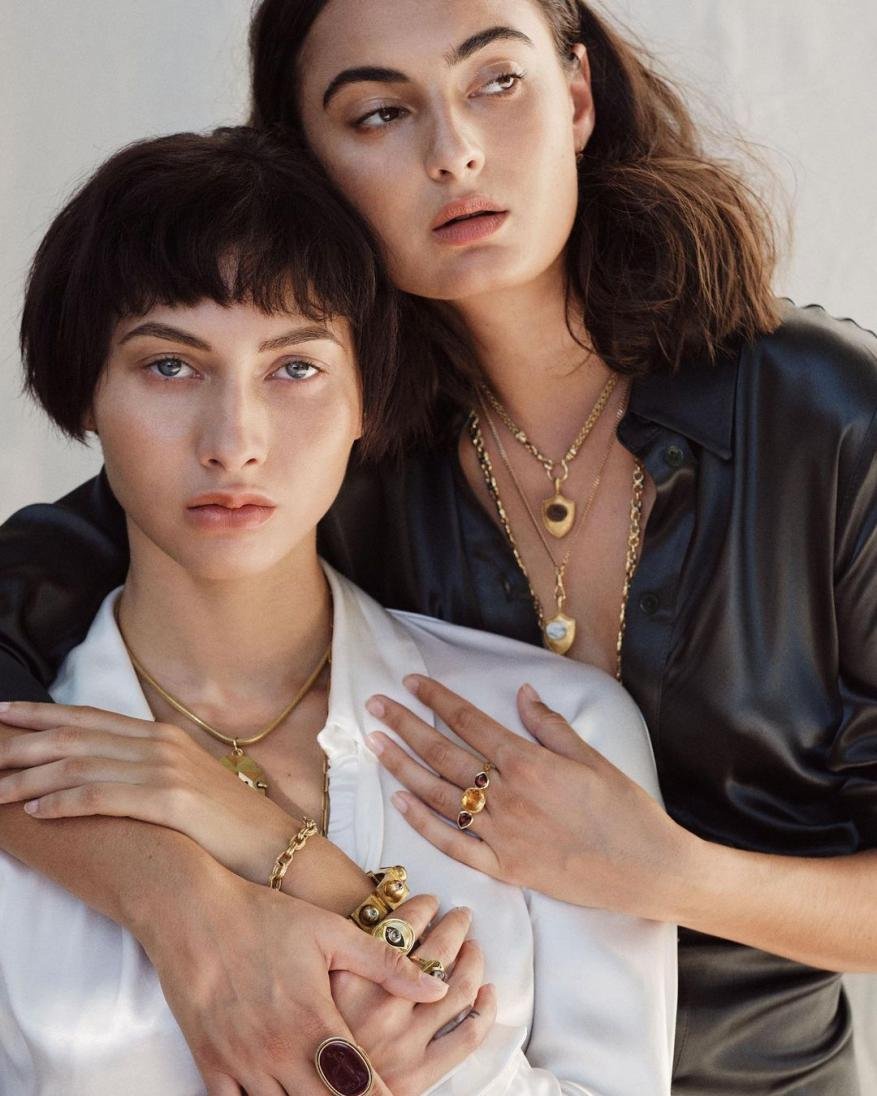 Two women pose in a studio wearing gold necklaces and rings, one in a white top, the other in a black top against a neutral background.