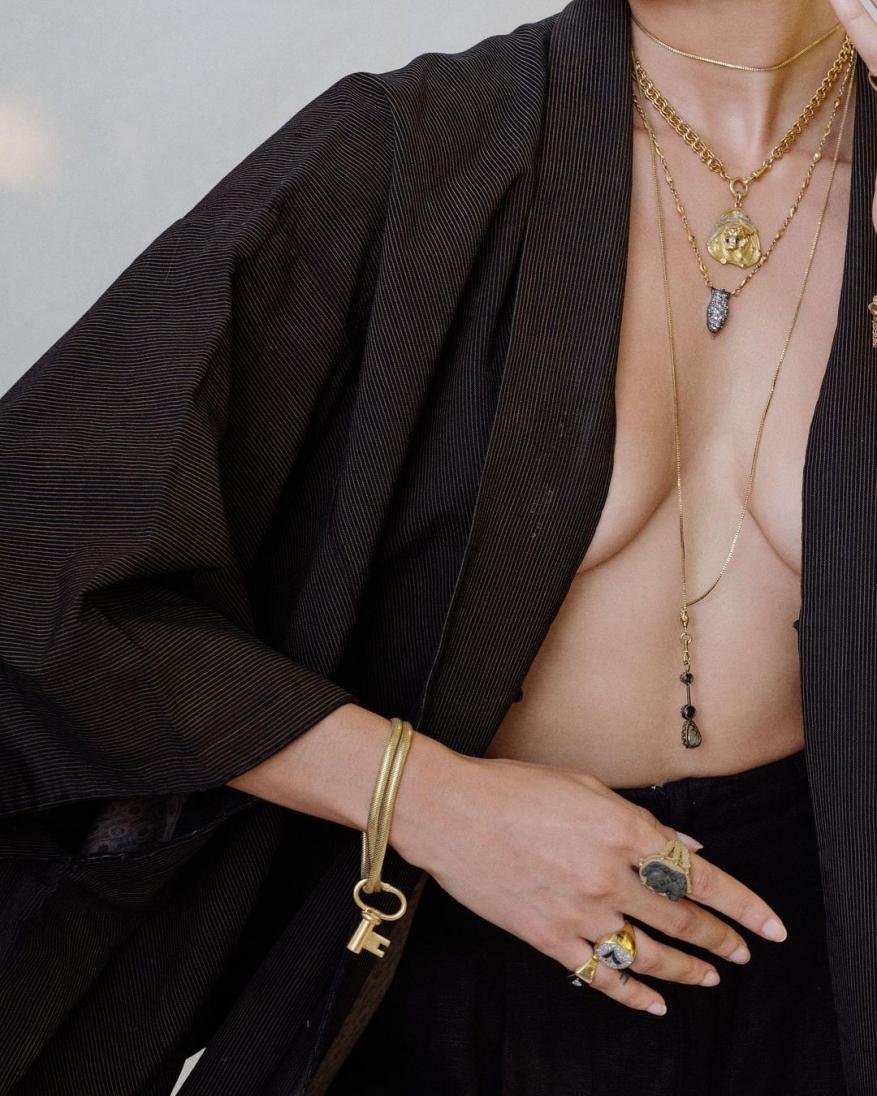 An adult female with several necklaces and rings poses indoors, wearing a black jacket with a gold key charm bracelet, against a white wall.