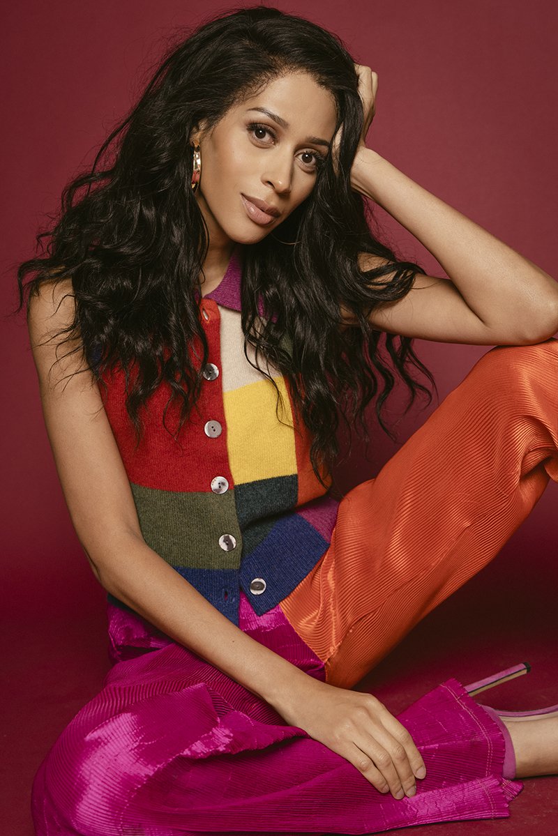 Attractive woman in a colorful sweater vest and pants sits in a photo studio, posing with hand near her head and relaxed smile.