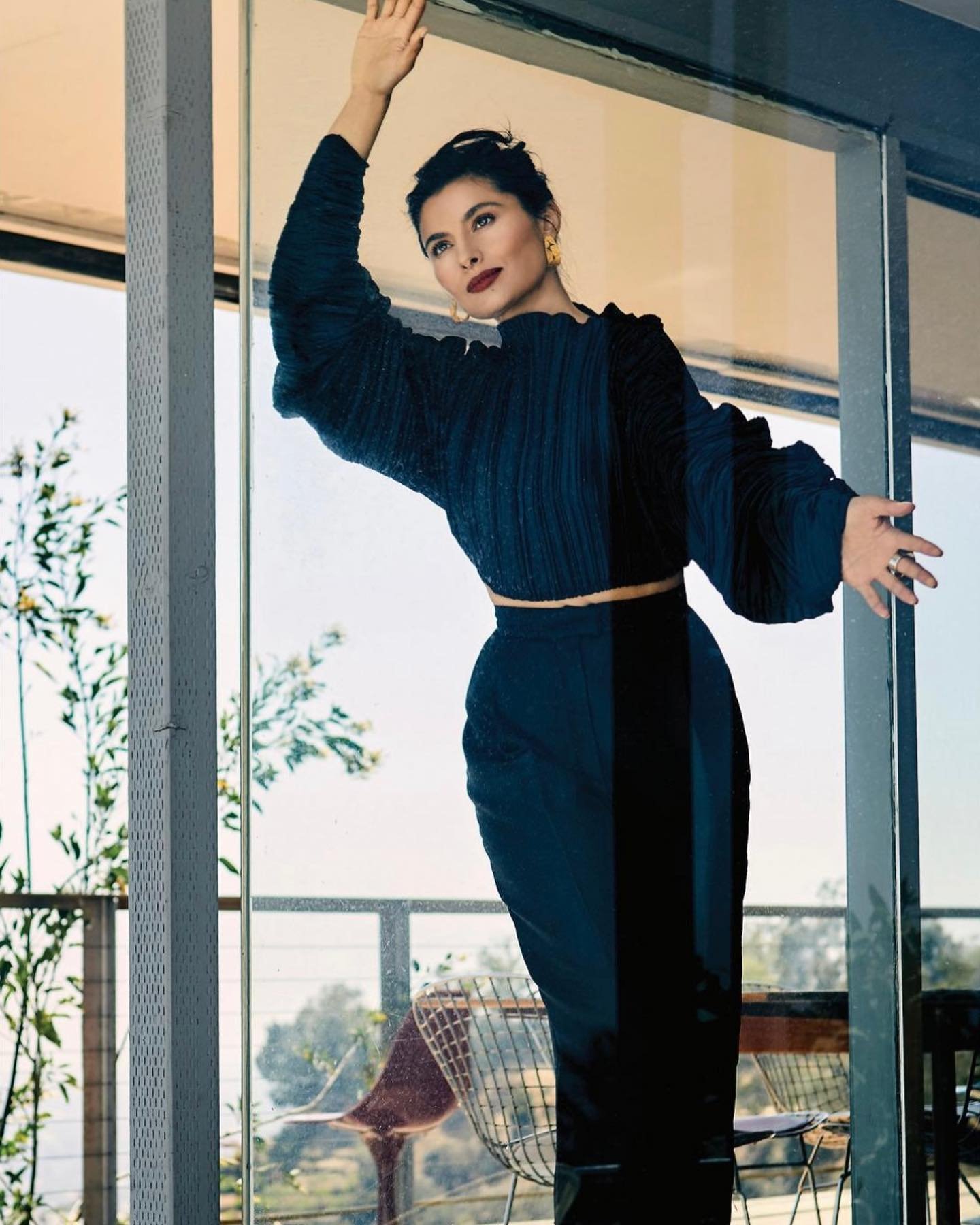 Elegant woman in stylish black separates poses by window with plants. Cropped top with long sleeves, high-waisted pants, jewelry, dark hair.