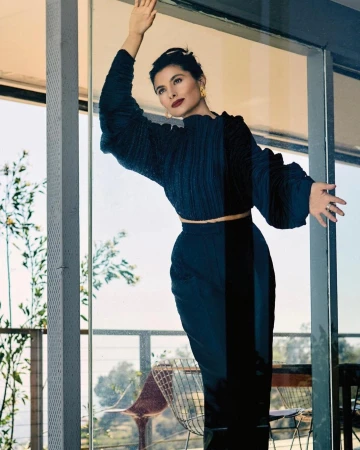 Elegant woman in stylish black separates poses by window with plants. Cropped top with long sleeves, high-waisted pants, jewelry, dark hair.