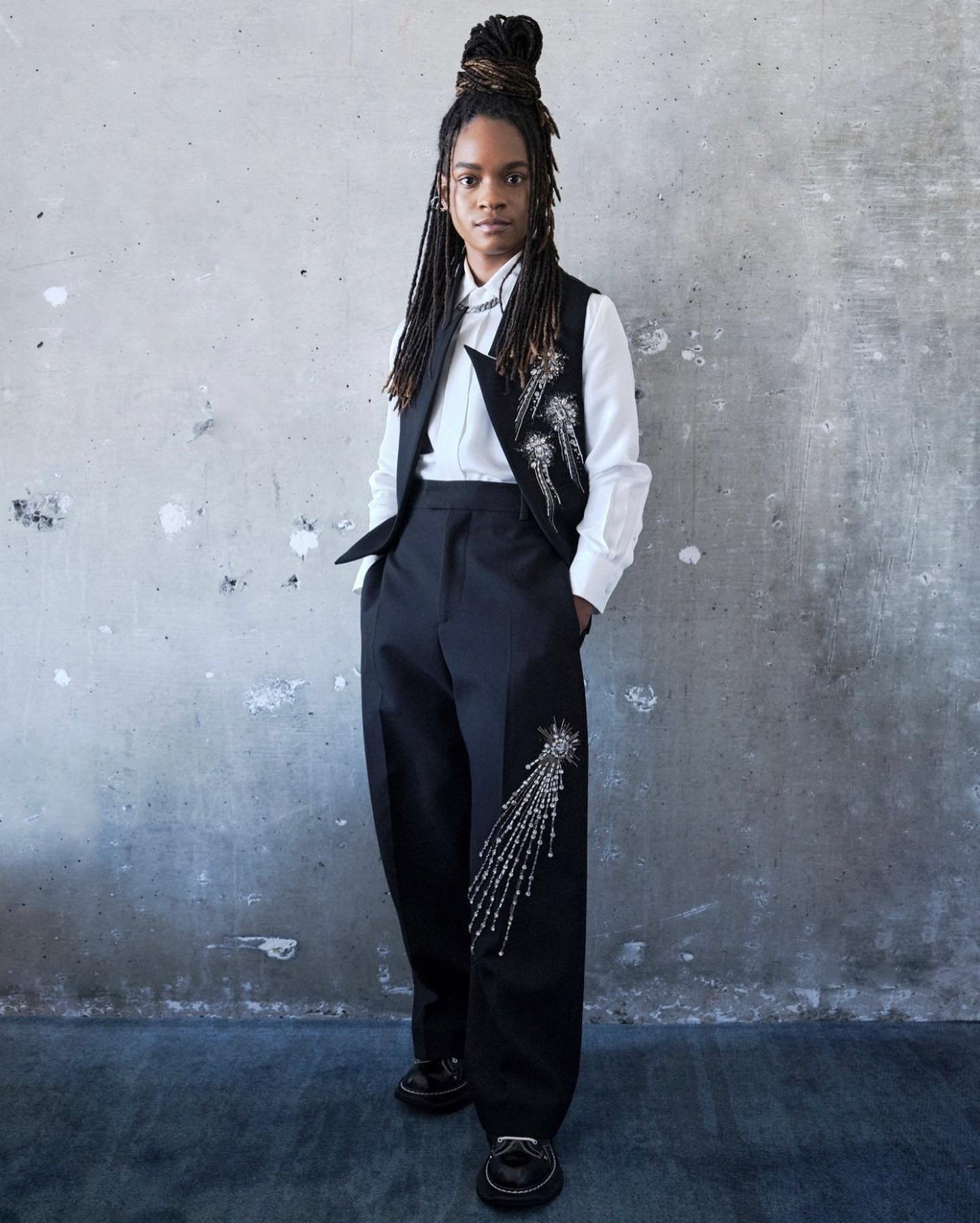 Portrait of an adult with dreadlocks wearing a black vest and trousers adorned with silver embellishments, standing against a concrete wall.