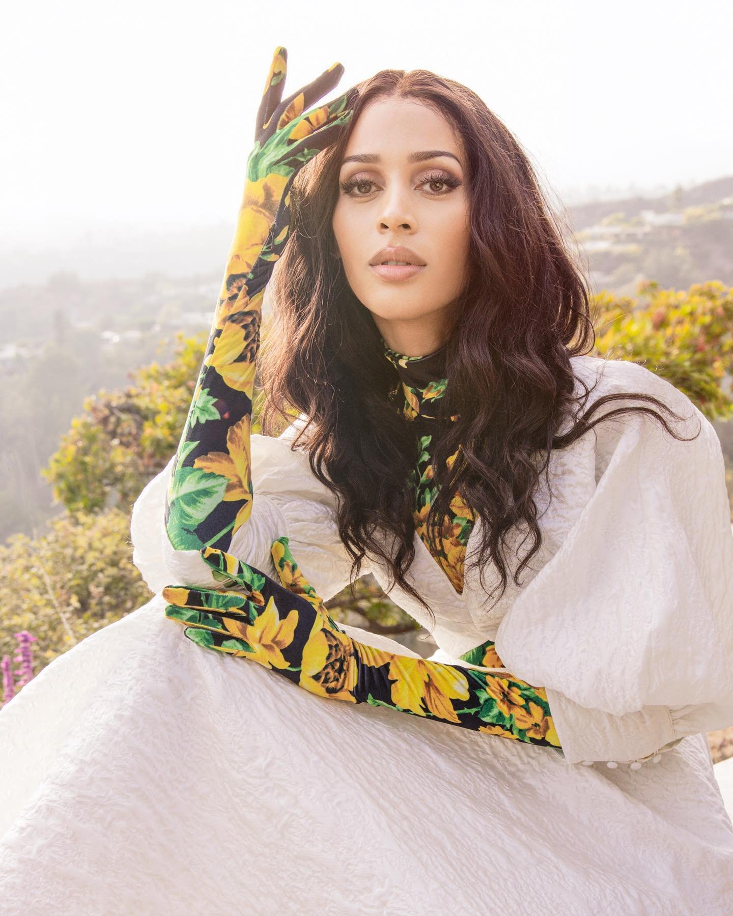 Portrait of a woman with dark hair wearing floral gloves and a white dress, posing outdoors with a blurred scenic background.