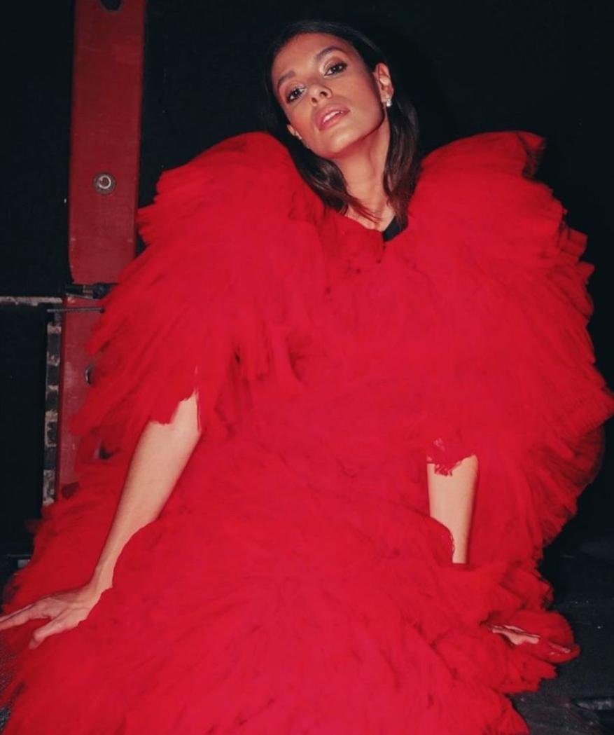 A woman with long brown hair poses in a voluminous red tulle dress against a dark background in a fashion or portrait style photo.