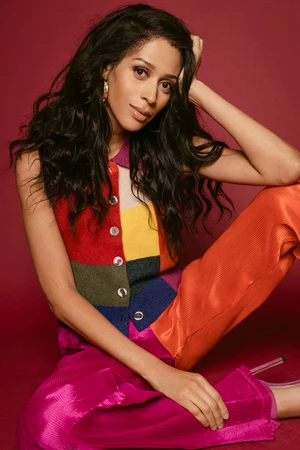 Woman with long dark hair wearing a multi-coloured knitted vest and bright trousers against a dark backdrop