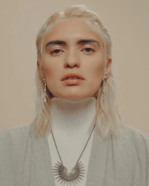 Close-up portrait of a blonde model wearing a white high-neck ribbed collar, grey blazer, and detailed pendant necklace.
