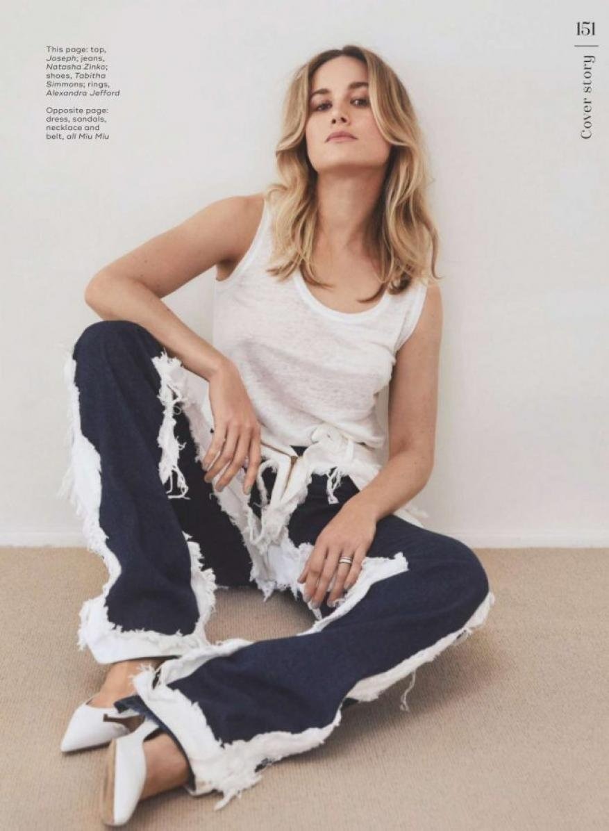 Woman with blonde hair sits on the floor in a white tank top and blue and white fringed pants, wearing white shoes in a studio.
