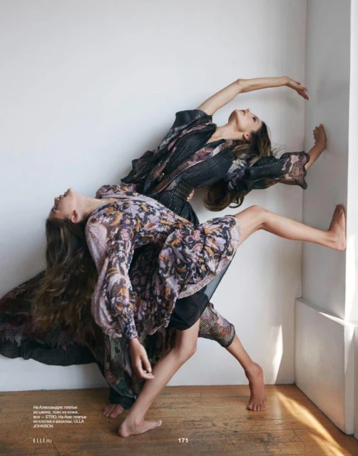 Two women in patterned dresses strike dynamic poses in front of a white wall, long hair flowing, barefoot, fashion editorial photography.