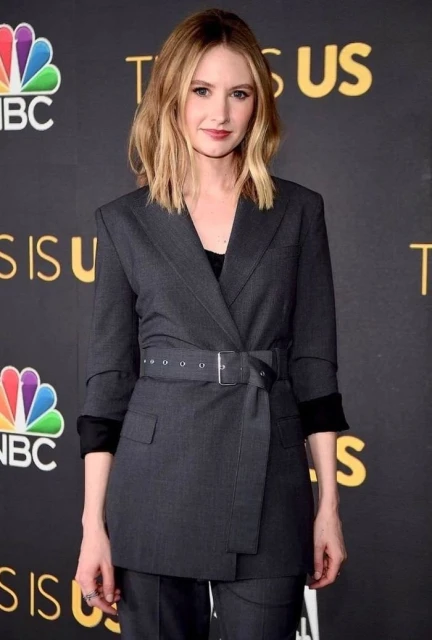 Woman with medium-length blonde hair poses on a red carpet wearing a belted grey pantsuit at a 'This Is Us' event.
