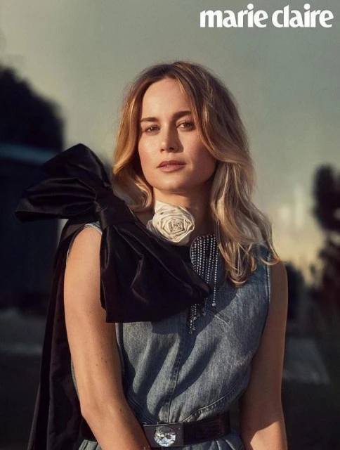 Elegant woman with blond hair wearing a denim dress with decorative embellishments and a black cape, posing in an outdoor setting.
