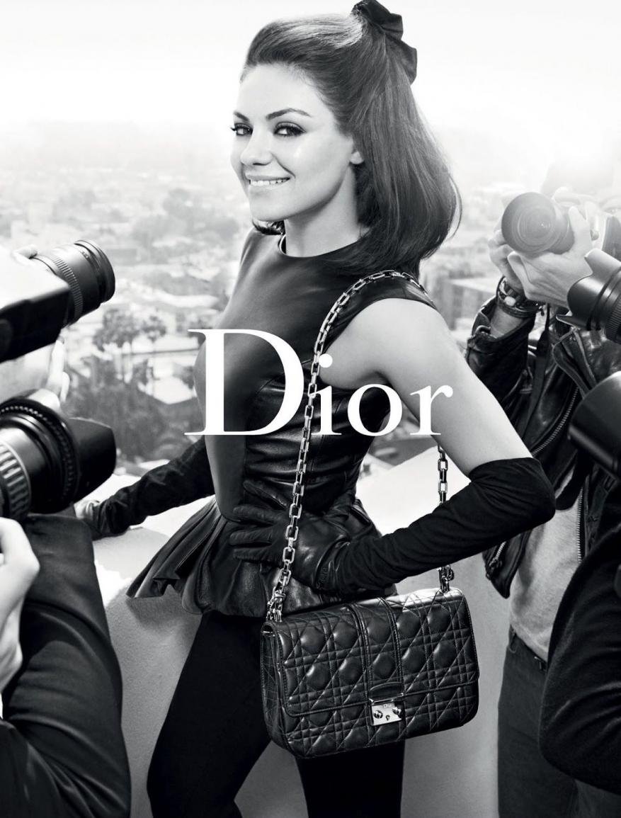 Mila Kunis poses with a Dior handbag, surrounded by photographers. The black-and-white image showcases a high-fashion aesthetic against a cityscape.