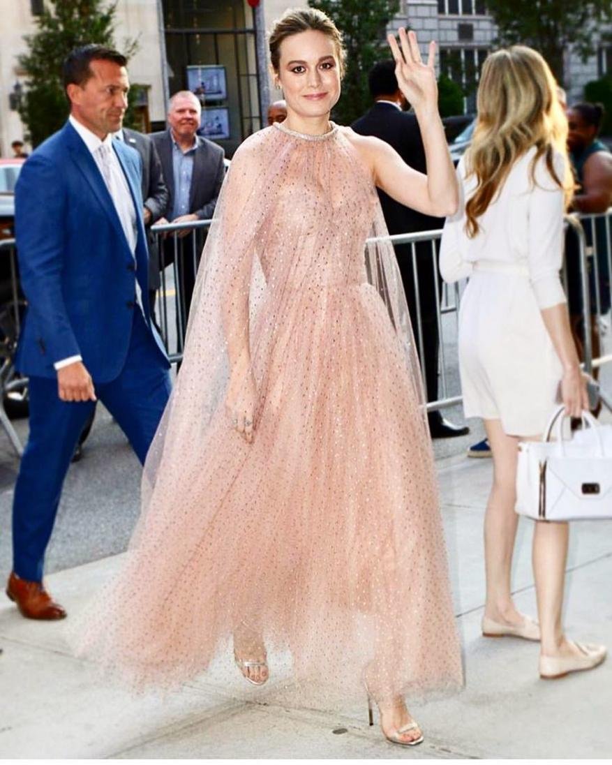 Brie Larson in a pink gown waves to the crowd at an outdoor event. The woman's dress is floor length with a cape detail.