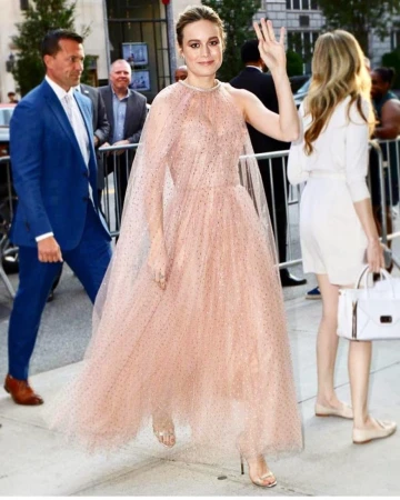 Brie Larson in a pink gown waves to the crowd at an outdoor event. The woman's dress is floor length with a cape detail.