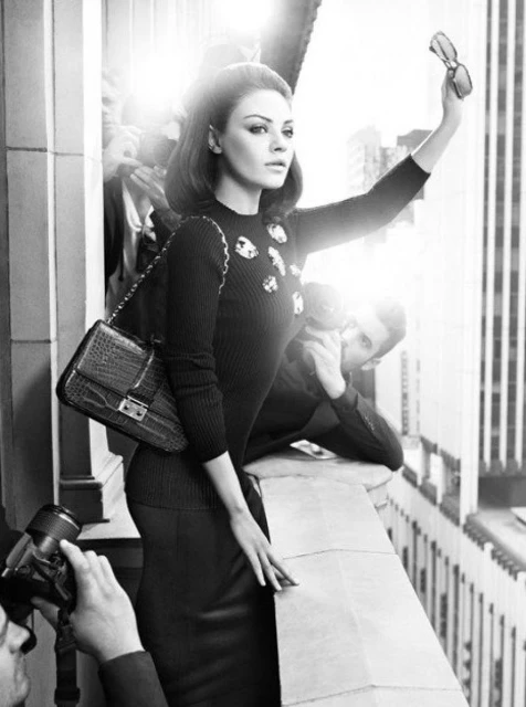 Black-and-white fashion shot of a brunette woman in a dark sweater, posing with photographers and a city skyline in the background.